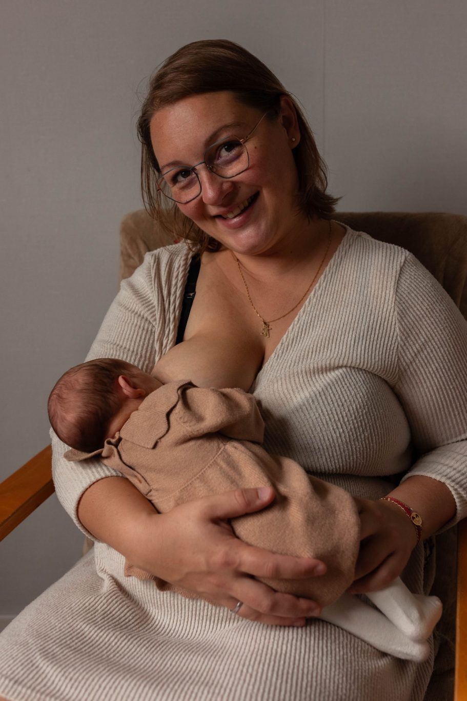 Femme souriante allaitant un bébé dans une chaise, vêtue d'un pull clair.